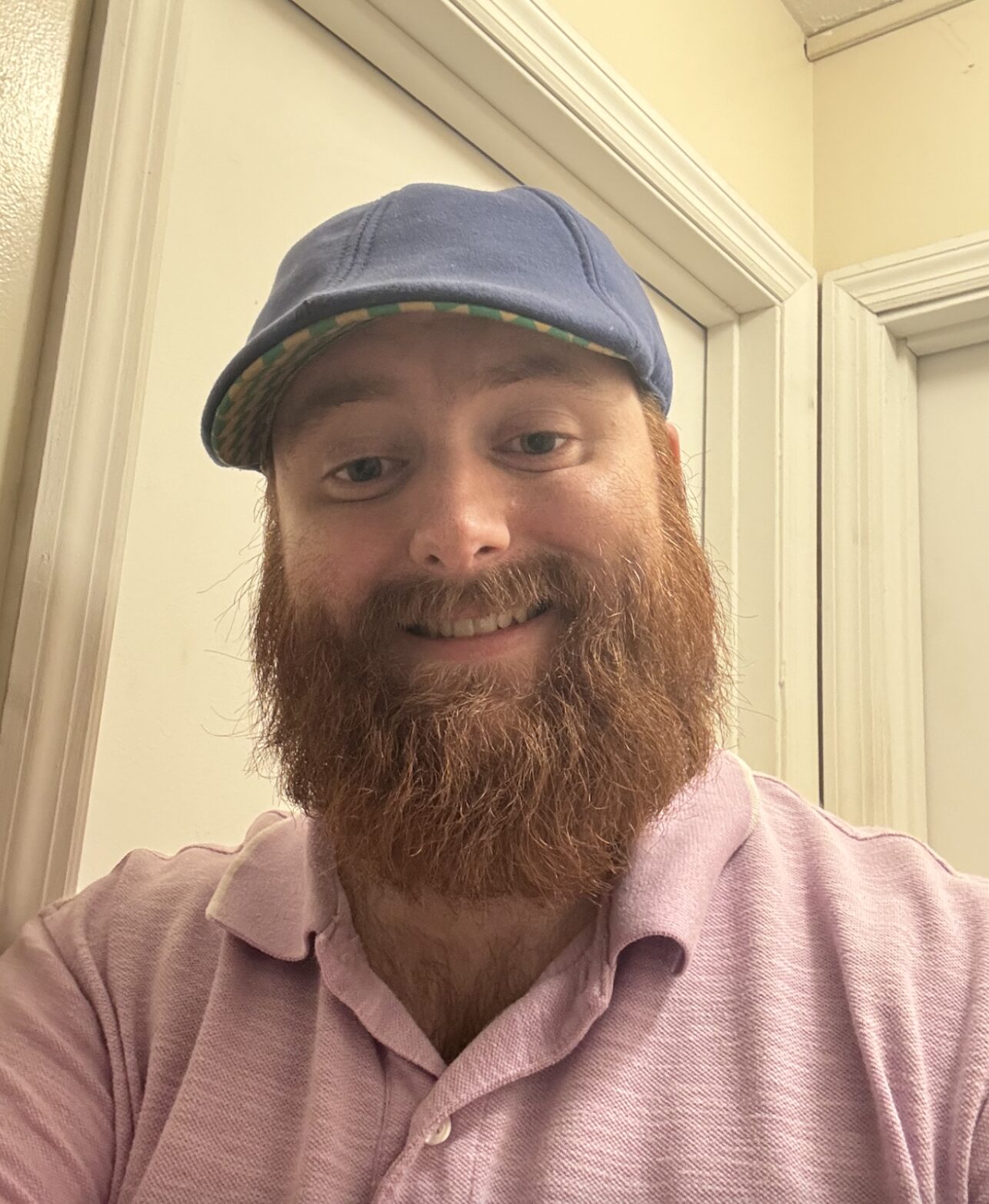 A smiling person with a red beard wears a blue cap and a light pink collared shirt, standing indoors in front of white doors.